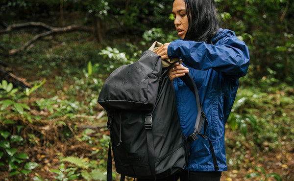 Woman wearing a blue raincoat pulling a hat out of the top pocket of the Shadow Guide; you can see rain drops beading off the fabric of the backpack.